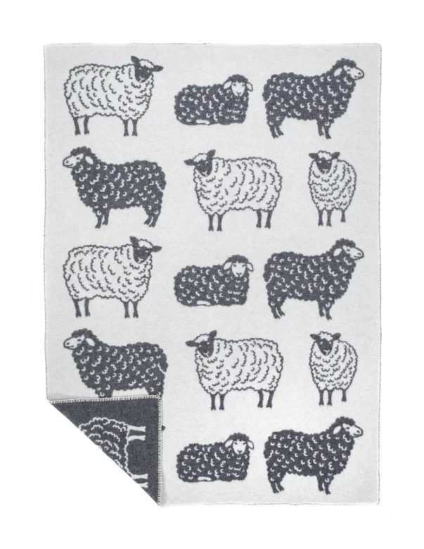 A wool blanket adorned with illustrations of black and white sheep on a gray background, featuring a fringe border, made by weaving mill Barker Textiles.