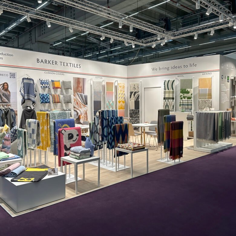 Barker Textiles booth at the 2024 Heimtextil trade fair displaying colorful blankets and cushion covers on racks and tables. Signs highlight materials like cotton, wool, and merino.